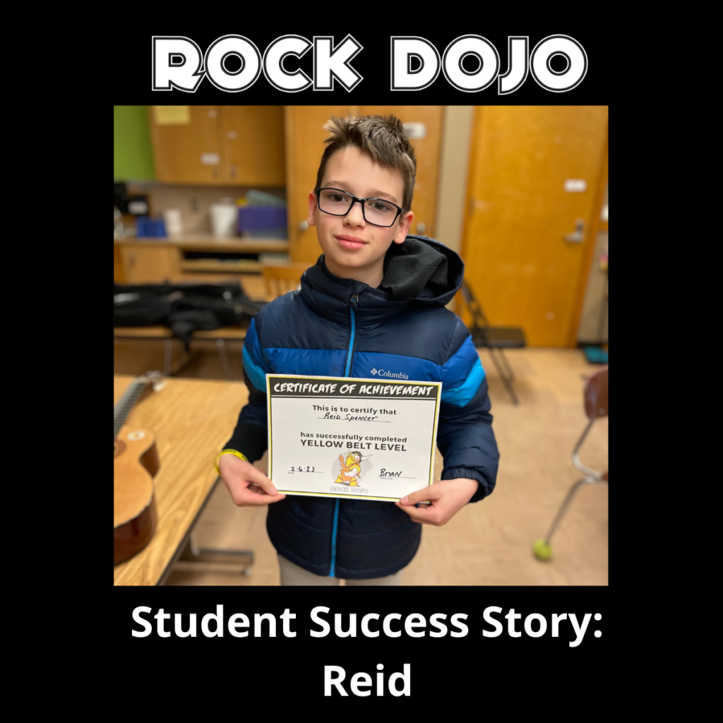 Reid with a big smile holding his yellow belt certificate in rock guitar from Rock Dojo.